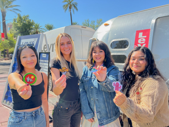 Four female college students posing in front of an airstream levi's bus holding up patches